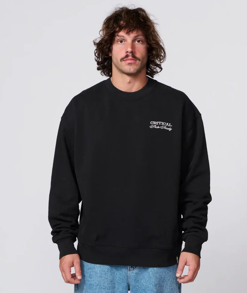 A man with curly brown hair and a mustache wearing a black crewneck sweatshirt with white 'CRITICAL Peanuts' text on the left chest, paired with light blue jeans, against a light gray background.